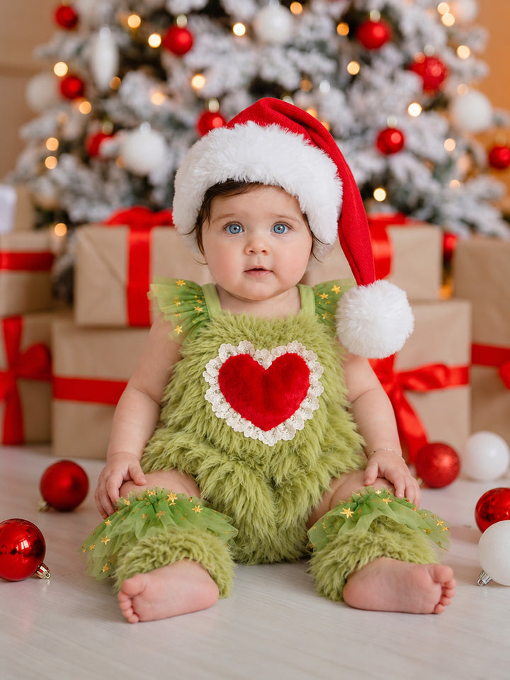 Baby sitting in front of a Christmas tree in the Little Grump green faux-fur romper with a red fur heart, matching leg warmers and a Santa hat - Before & Ever Kids