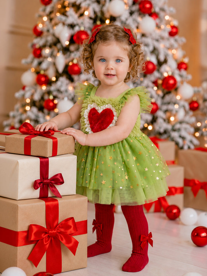 Girl standing beside wrapped presents in the Miss Grump green tulle Christmas dress with a red fur heart and gold stars, worn with red bow tights and red hair bows - Before & Ever Kids