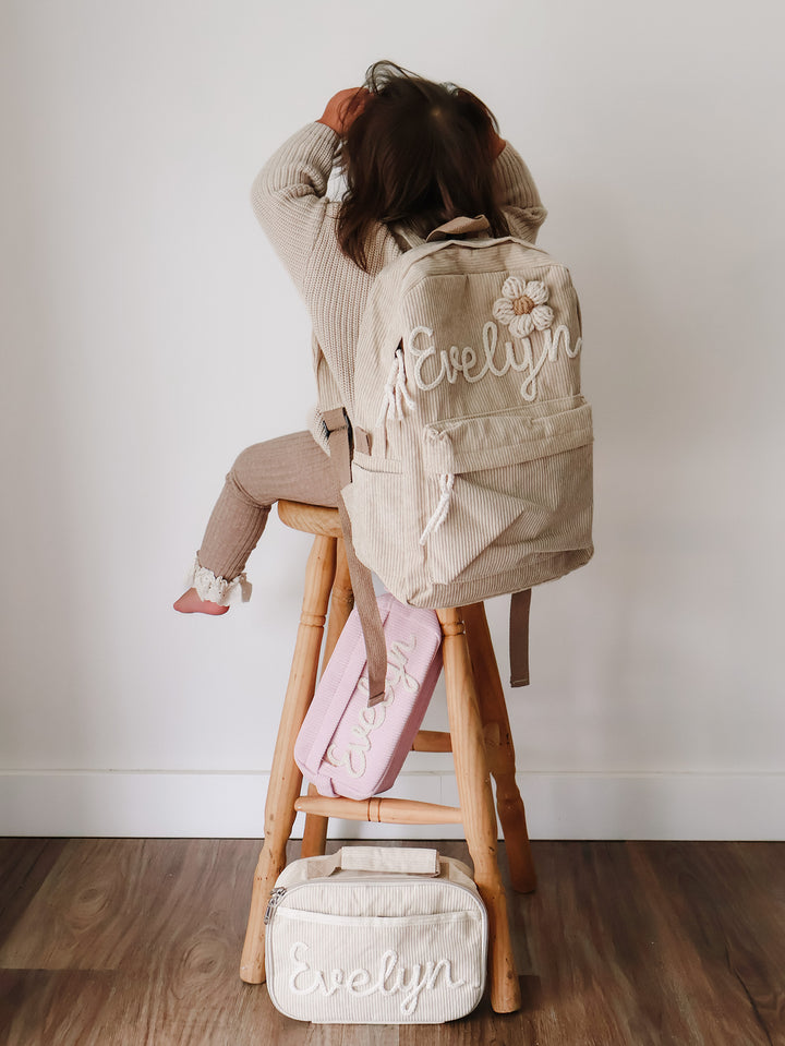 Beige backpack embroidered Evelyn worn by a girl on a stool, matching lunch bag and pink pouch on the floor beneath