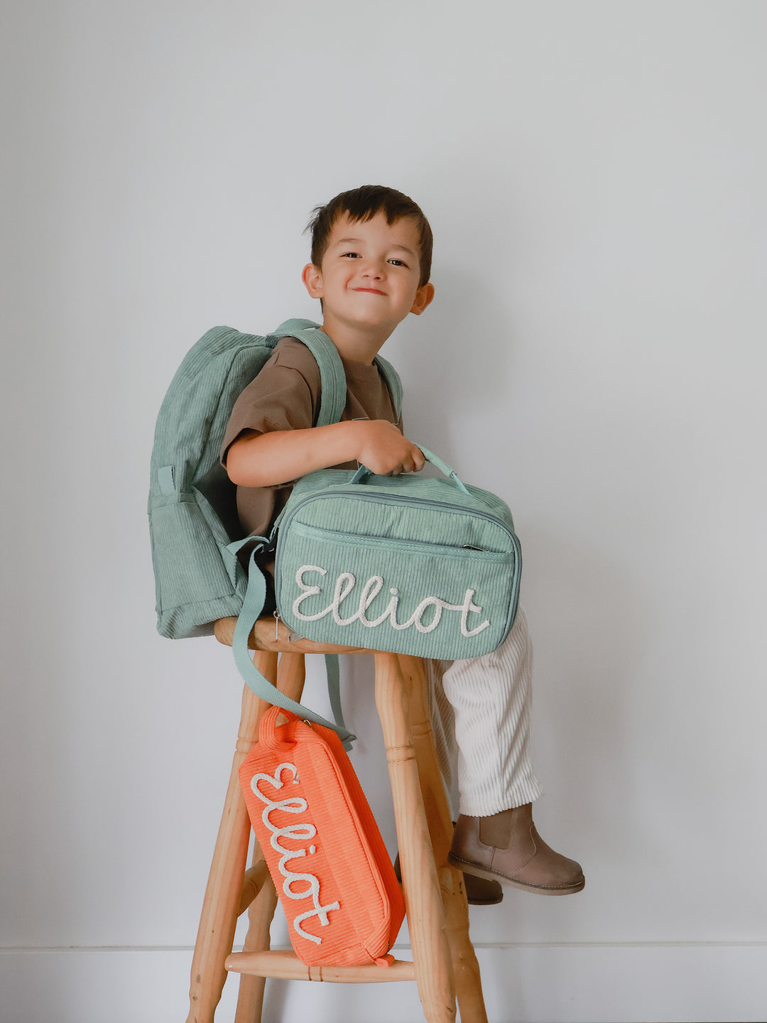 Sage green backpack and lunch bag embroidered Elliot, worn and held by a smiling boy, orange pouch on the stool below