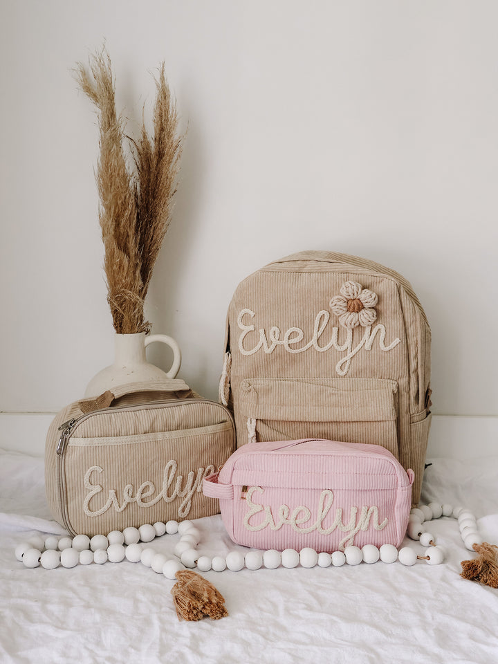 Beige lunch bag and backpack with a pink pouch, all embroidered Evelyn, styled together on a bed