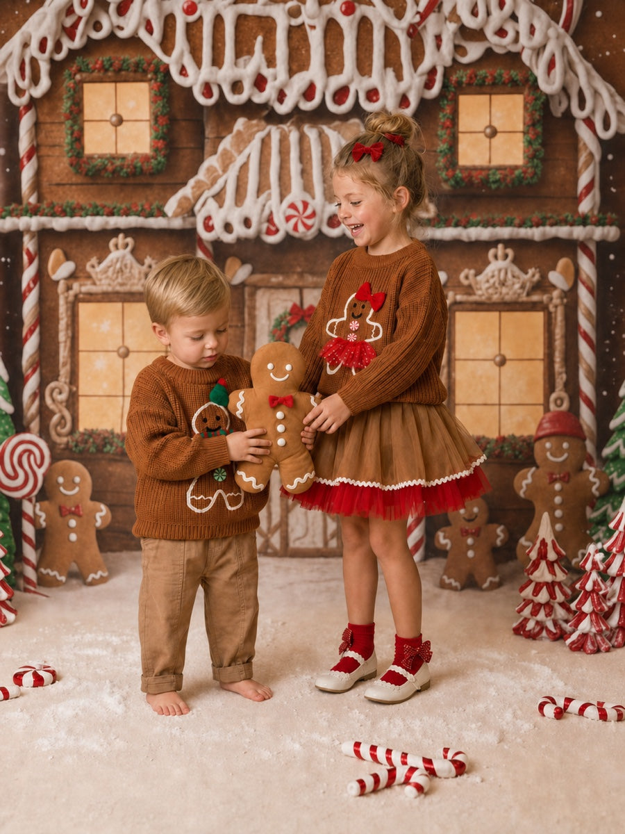Mr and Miss Gingerbread sweaters on a boy and a girl at a gingerbread house, the boy in the Mr Gingerbread caramel knit looking down at the gingerbread man plush he is holding, the girl laughing in the Miss Gingerbread knit over her Sugar and Spice caramel and red tulle skirt - Before & Ever Kids