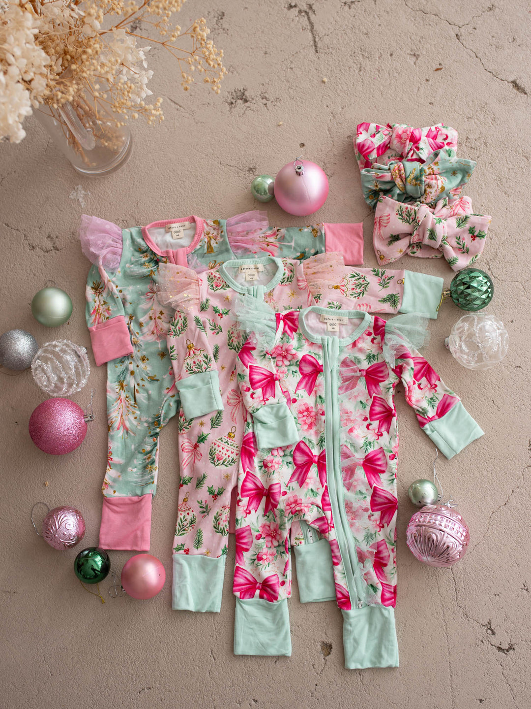 All I Wish Christmas baby onesies and topknots laid out together including the Pink Holly print - Before & Ever Kids