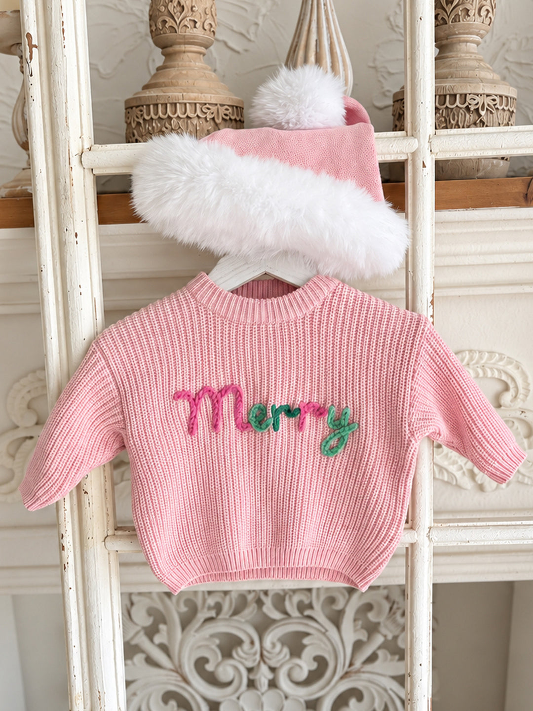 Baby pink knitted Santa hat with white fluffy trim and pom-pom, draped over a hanging pink Merry knit sweater