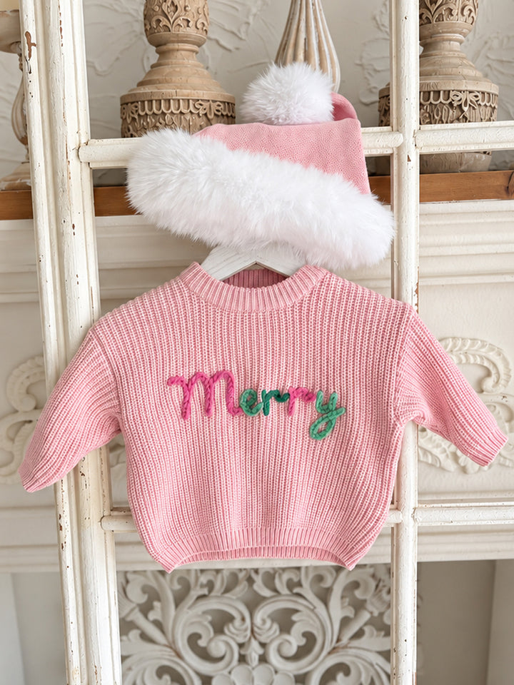 Baby pink knitted Santa hat with white fluffy trim and pom-pom, draped over a hanging pink Merry knit sweater