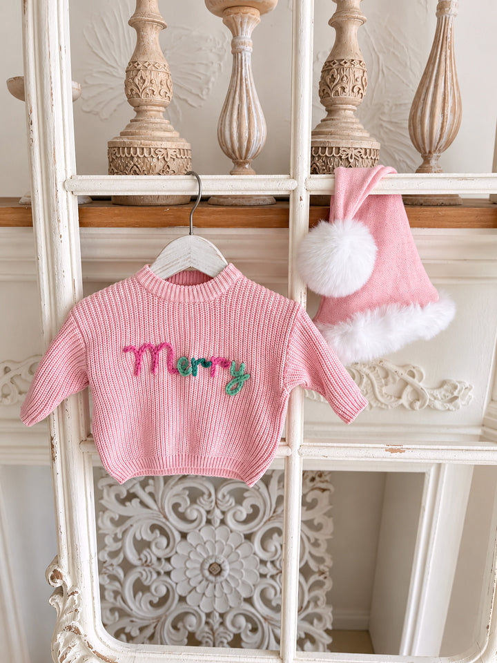 Baby pink knitted Santa hat with white fluffy trim and pom-pom, hanging beside a pink Merry knit sweater