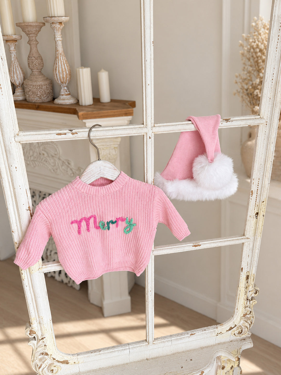 Baby pink knitted Santa hat with white fluffy trim and pom-pom, hanging on a vintage window frame beside a pink Merry knit sweater