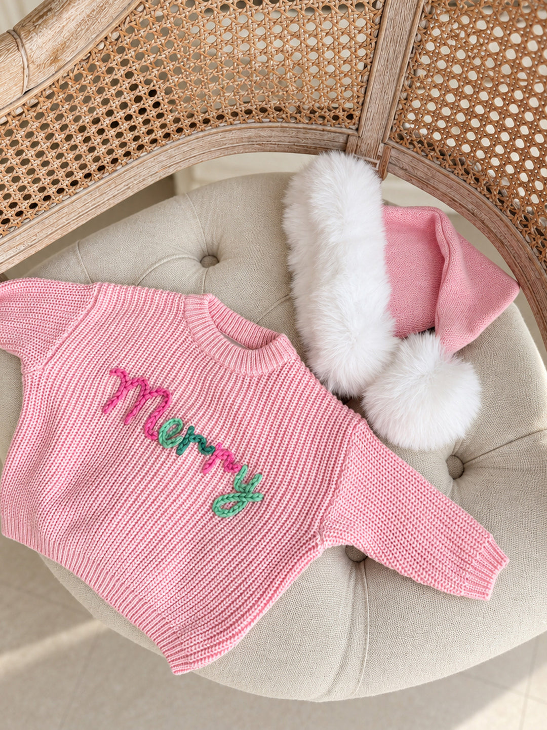 Baby pink knitted Santa hat with white fluffy trim and pom-pom, flat lay with a pink Merry knit sweater