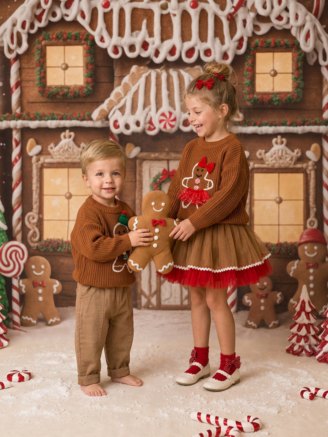 Toddler boy in the Mr Gingerbread knitted sweater with a gingerbread man motif and green beanie, standing with a girl in the Miss Gingerbread sweater over the Sugar and Spice caramel and red tulle skirt, holding a gingerbread man plush - Before & Ever Kids