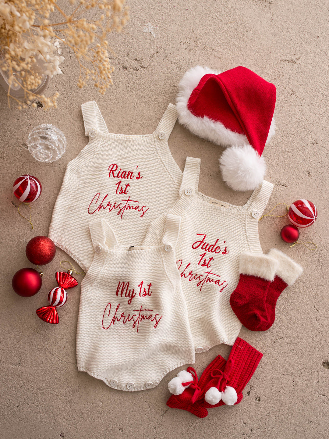 Santa Baby Knitted Booties in red cotton knit with white pom poms, beside three knitted rompers and a Santa hat - Before & Ever Kids