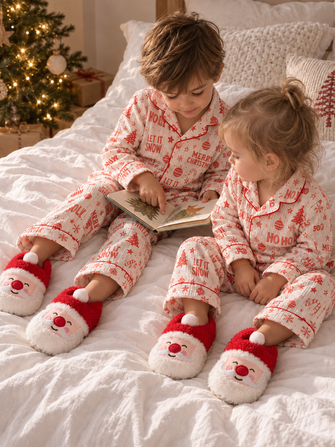 Santa Baby Slippers red plush Santa face slippers worn by two children reading a book on a bed at Christmas - Before & Ever Kids