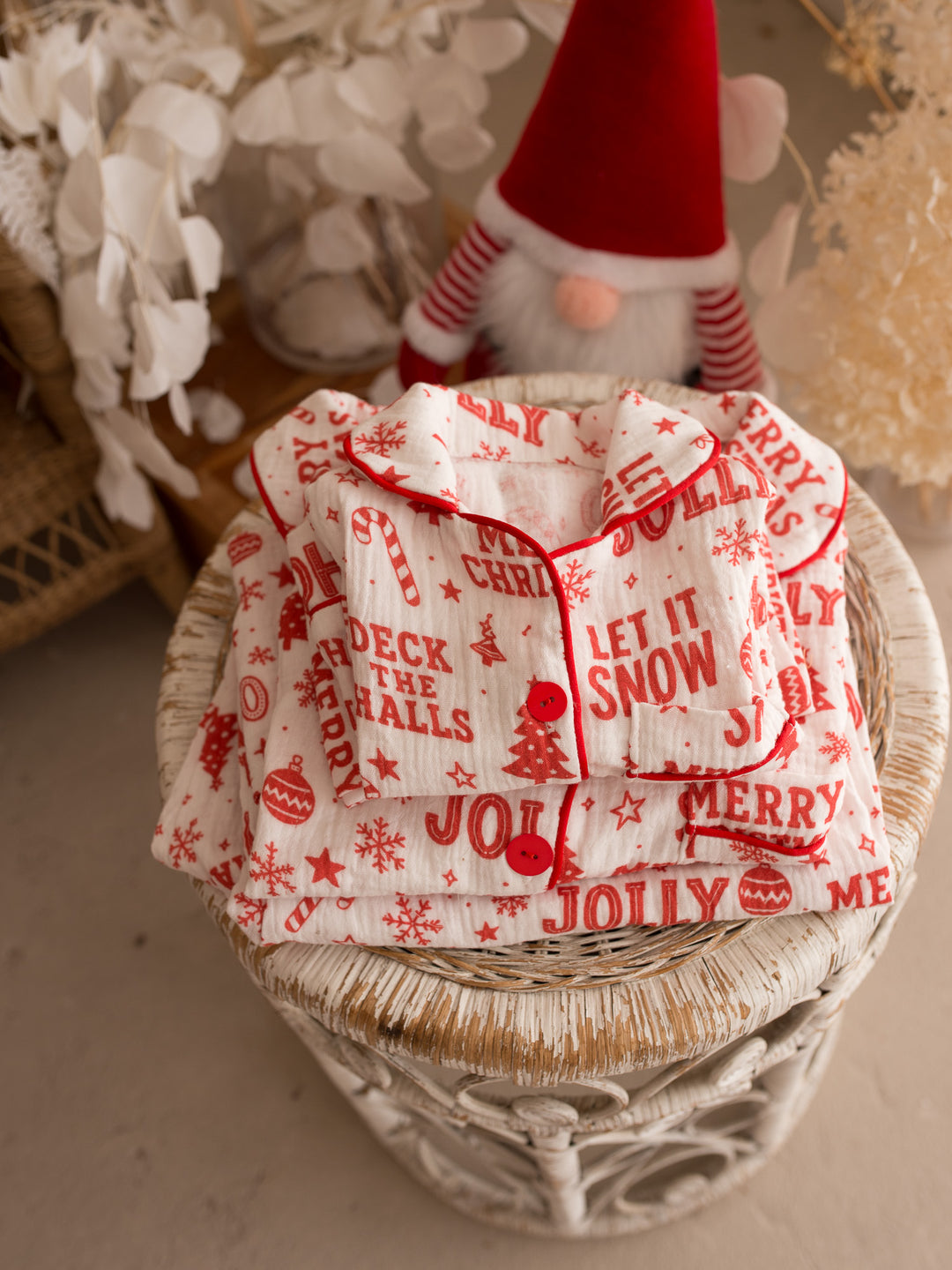 Twas the Night family Christmas pyjamas, white cotton muslin with a red Christmas print, sleep shirt and pants folded on a wicker stool beside a Christmas gnome - Before & Ever Kids