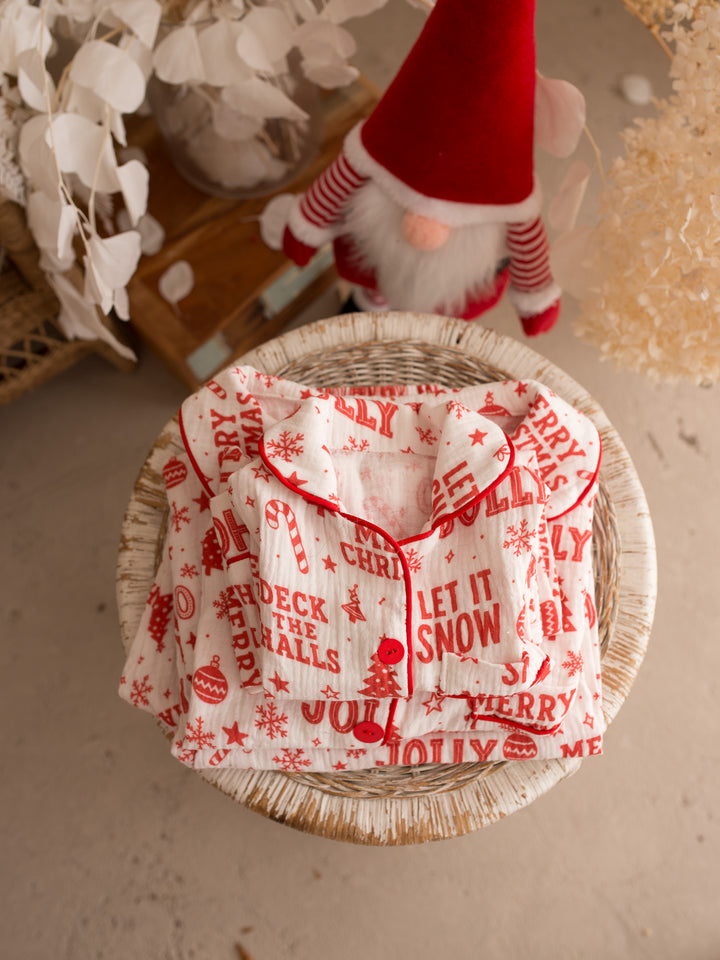 Twas the Night family Christmas pyjamas, white cotton muslin with a red Christmas print, the folded set on a wicker stool, close up - Before & Ever Kids