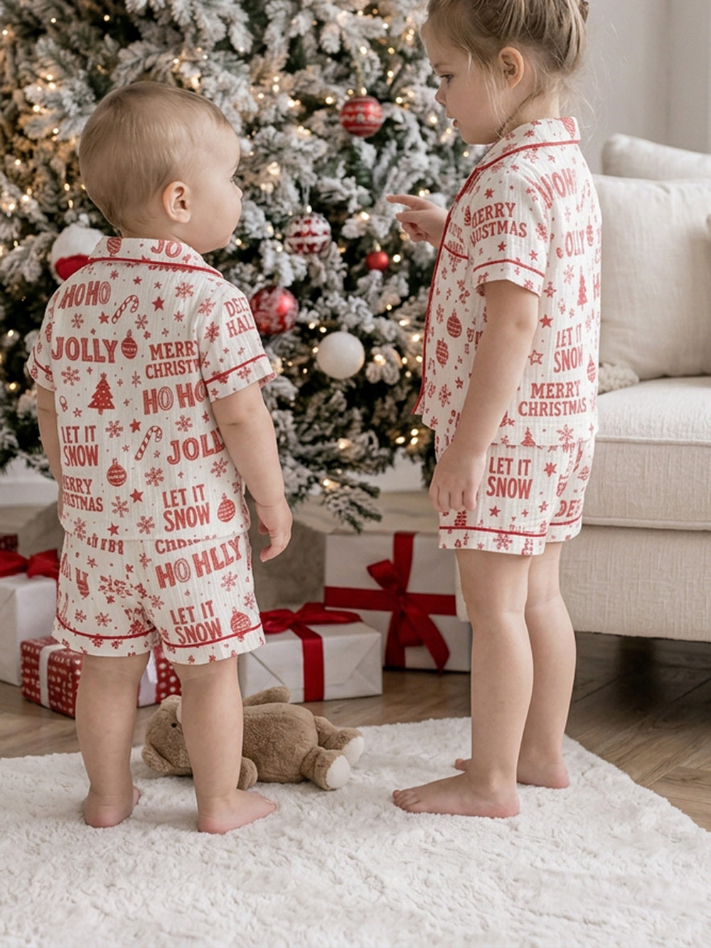 Twas the Night family Christmas pyjamas, white cotton muslin with a red Christmas print, baby and the girl in short sleeve shirts and shorts standing by the tree with a teddy - Before & Ever Kids