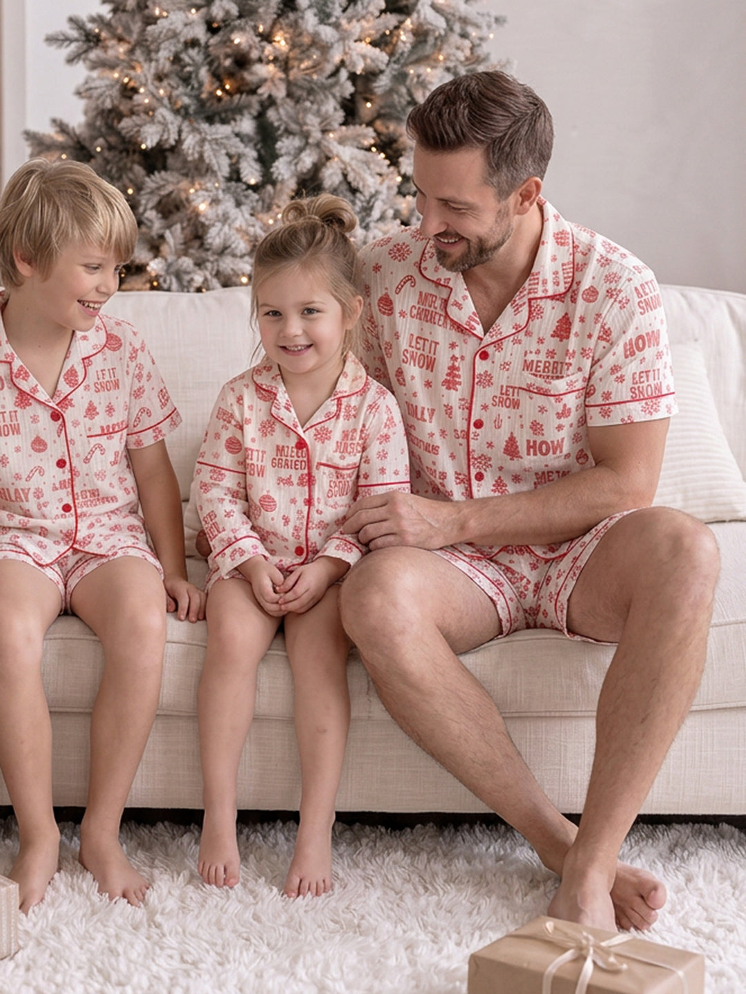 Twas the Night family Christmas pyjamas, white cotton muslin with a red Christmas print, dad in the short sleeve shirt and shorts on the sofa with the boy and girl - Before & Ever Kids