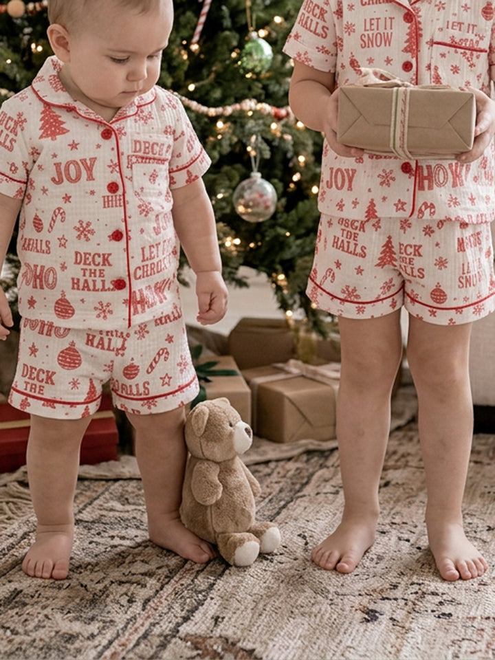 Twas the Night family Christmas pyjamas, white cotton muslin with a red Christmas print, close crop of the baby in shorts standing with a teddy by the presents - Before & Ever Kids
